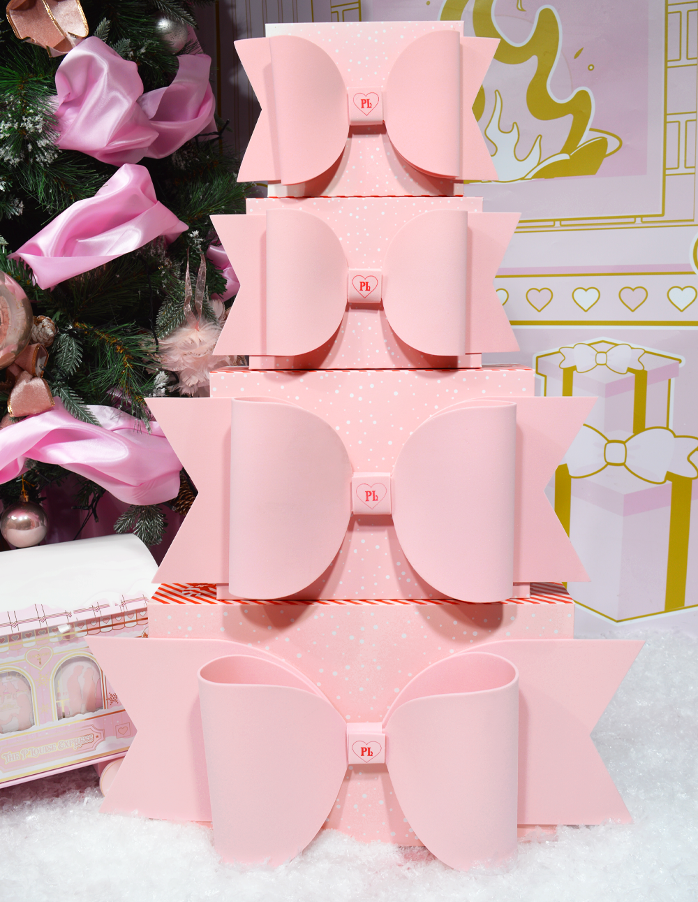 Pila de cajas de regalo rosadas de P.Louise con lazos exhibidas sobre un fondo de temática navideña con adornos.