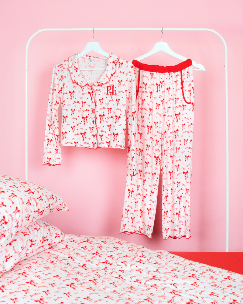 Conjunto de pantalones de pijama con lazo navideño para niñas de P.Louise, que presenta un diseño de lazo rojo sobre rayas rosadas y blancas colgadas de perchas en un dormitorio rosa con ropa de cama a juego.