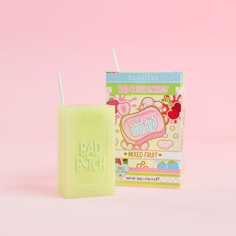 Et grønt stykke sæbe med 'Bad Bitch' indgraveret på det, ledsaget af en farverig juicebokspakke med 'Mixed Fruit' mærket på, præsenteret på en lyserød baggrund.