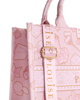 Un primer plano de un bolso tote rosa claro con un patrón dorado de postres y el texto vertical 'P.LOUISE' en una correa central, con un cierre de hebilla dorada.