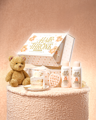 An image showing a fluffy table with a P.Louise Hair To The Throne haircare set showing a shampoo and conditioner in coconut cake scents and a scalp massager with a teddy bear and a slice of cake next to it
