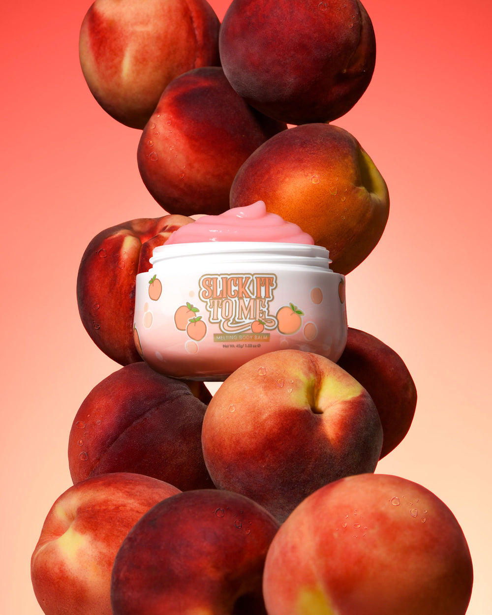 Tarro de P. Louise Slick It To Me Peach Melting Body Balm con crema rosa rodeada de melocotones sobre un fondo degradado