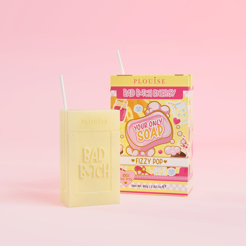Una pastilla de jabón amarilla con la inscripción 'Bad Bitch' grabada, acompañada de una colorida caja de jugo con la etiqueta 'Fizzy Pop', presentada sobre un fondo rosa.