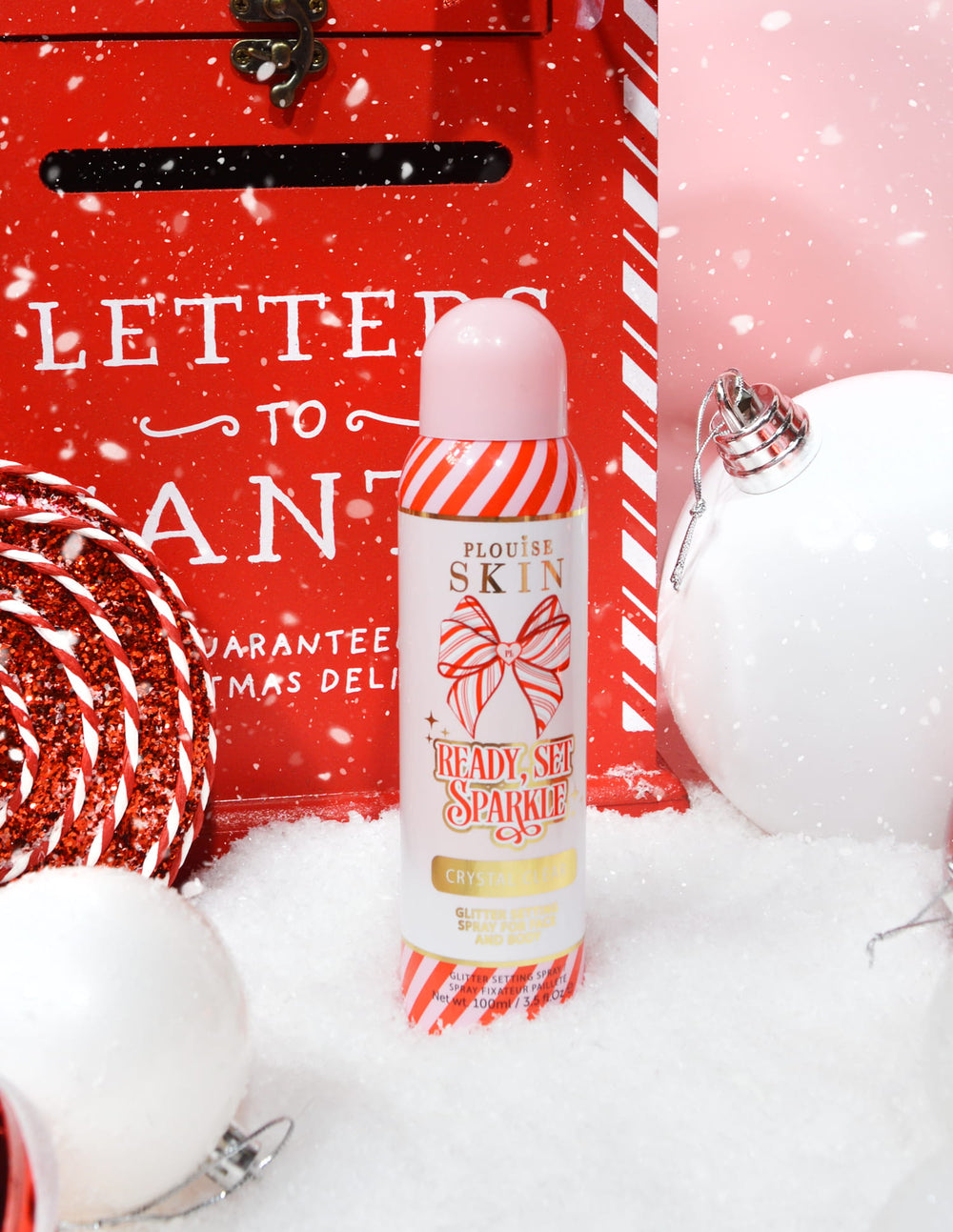 Una botella de spray fijador de purpurina navideña P. Louise Ready Set Sparkle rodeada de adornos festivos como nieve, adornos y un buzón navideño.
